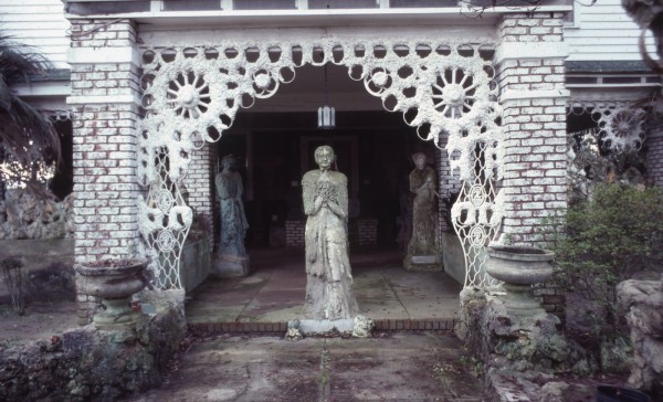 A concrete sculpture of a figure stands underneath an ornate, white entry way