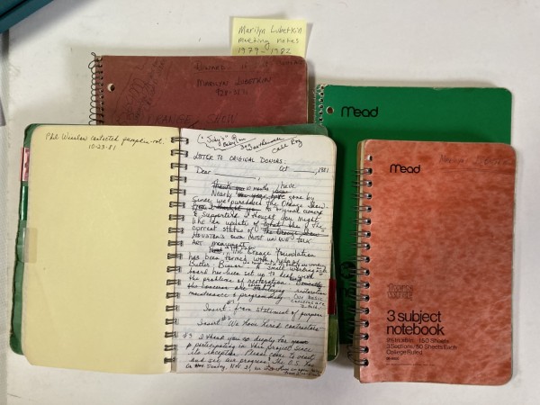 Several worn notebooks on a table with one open to a page with a handwritten letter draft to donors by Marilyn Oshman.