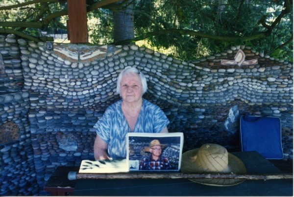 a woman sits at a table in front of a mosaicked wall. On the table is a picture of a man with a cowboy hat.