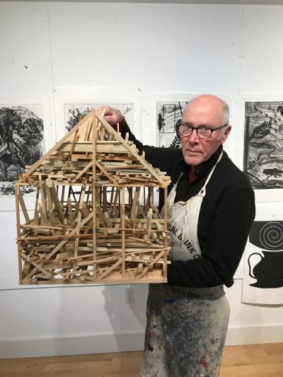 Gregg Blasdel stands in a gallery with black and white photography behind him. He is holding a sculpture in the shape of a house made of wooden sticks.