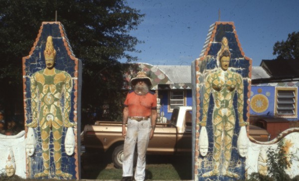 A person in a red shirt and white pants, Eddie Owens Martin, stands between two decorative pillars with colorful bas reliefs of figures. A car and the rest of Pasaquan is behind him. . 